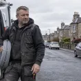 A tradesperson carrying a coil of cable from their van along a street of sandstone houses